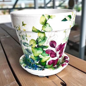 May include: White ceramic flower pot with a matching saucer, decorated with green, blue, and purple botanical illustrations. The pot has a wide rim and a slightly tapered shape, suitable for indoor or outdoor use.