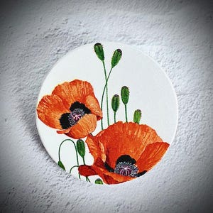 Peut inclure: Un objet décoratif rond et blanc représentant une peinture à l'aquarelle de deux coquelicots orange vif avec des centres noirs et des tiges et boutons verts. Le motif floral est posé sur un fond blanc.