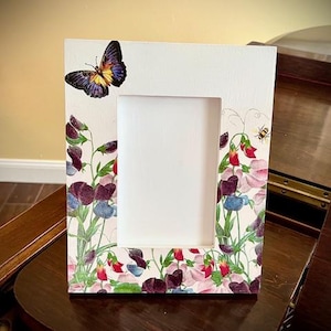 May include: White rectangular picture frame with a floral design featuring purple, red, and pink flowers, a butterfly, and a bee. The frame is decorated with a botanical illustration.