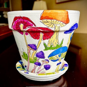 Puede incluir: Una maceta de cerámica blanca con un diseño de setas de colores. La maceta tiene un platillo y está decorada con setas rojas, naranjas, amarillas, verdes, azules y moradas.