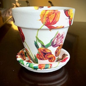 May include: White ceramic flower pot with a saucer. The pot and saucer are decorated with a floral pattern of tulips and butterflies in shades of orange, red, pink, yellow, and green.
