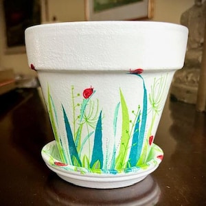 Puede incluir: Maceta de terracota blanca con un diseño pintado a mano de hierba verde y azul y mariquitas rojas. La maceta tiene un platillo a juego.