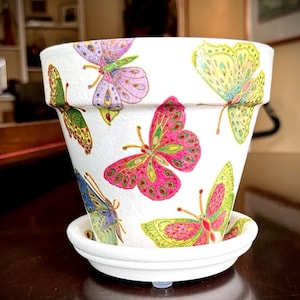 May include: White ceramic flower pot with a butterfly design. The butterflies are colorful and have intricate details. The pot has a matching saucer.