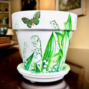 Puede incluir: Maceta de cerámica blanca con platillo a juego. La maceta está decorada con una mariposa verde e ilustraciones de lirios de los valles. La maceta tiene una superficie texturizada y está diseñada para uso en interiores o exteriores.