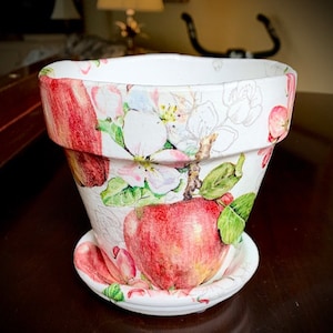 Puede incluir: Una maceta de cerámica blanca con un diseño floral que presenta flores rosas y blancas, hojas verdes y manzanas rojas. La maceta tiene un platillo a juego.