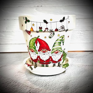 May include: White ceramic planter with a Christmas gnome theme. The planter features three gnomes wearing red and green hats and holding presents. The planter is decorated with a string of lights, stars, and other Christmas decorations.