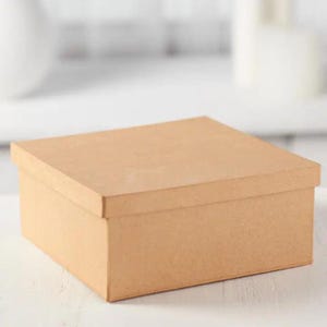 Op de afbeelding: Een vierkante, lichtbruine kartonnen doos met een bijpassend deksel. De doos heeft een gladde, matte afwerking en een eenvoudige, strakke constructie met rechte randen en hoeken, wat een neutrale uitstraling geeft voor opslagdoeleinden.