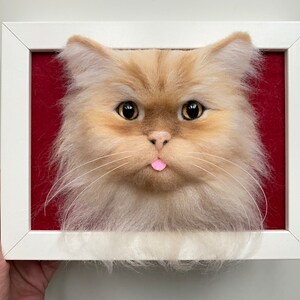 May include: A needle felted cat head portrait in a white frame. The cat has light brown fur, a pink tongue sticking out, and large brown eyes.