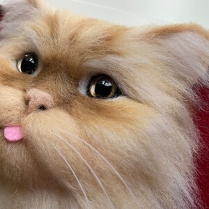 May include: Close-up of a needle felted cat head with a pink tongue sticking out. The cat has brown and white fur and large, dark eyes.