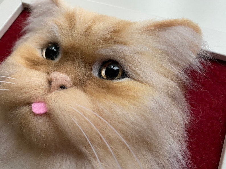 May include: Close-up of a needle felted cat head with a pink tongue sticking out. The cat has brown and white fur and large, dark eyes.