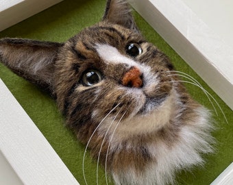 May include: A framed needle felted portrait of a tabby cat with white markings. The cat has brown and white fur and is looking directly at the viewer. The frame is white with a green background.
