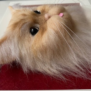 May include: Close-up of a needle felted cat head with white and light brown fur. The cat has a pink tongue sticking out and is looking to the right.