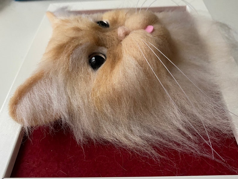 May include: Close-up of a needle felted cat head with white and light brown fur. The cat has a pink tongue sticking out and is looking to the right.