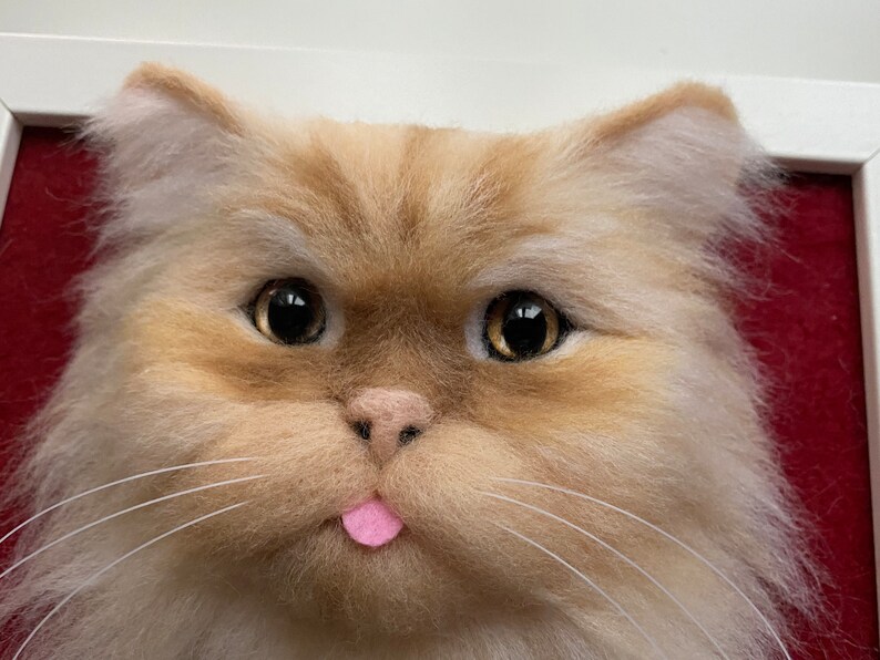 May include: Close-up of a fluffy, light brown and white cat with its tongue sticking out. The cat has large, dark eyes and a pink nose. The cat is framed in a red mat.