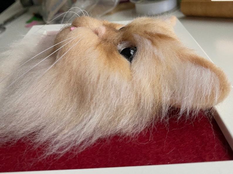 May include: Close-up of a needle felted cat head with a light brown and white fur. The cat has a pink nose and one black eye.