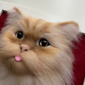 May include: Close-up of a felted cat head with a pink tongue sticking out. The cat has brown fur with white highlights and large brown eyes.