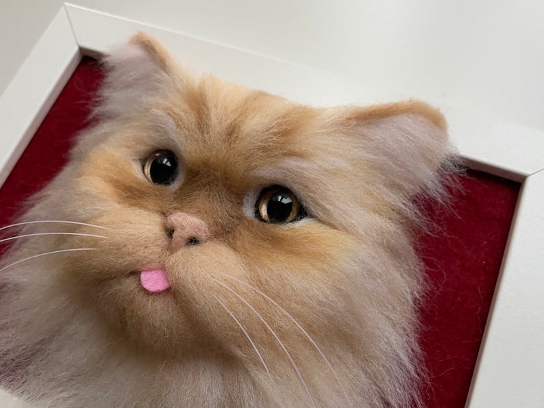 May include: Close-up of a felted cat head with a pink tongue sticking out. The cat has brown fur with white highlights and large brown eyes.