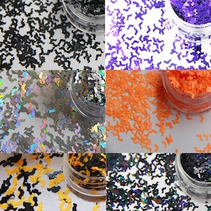 Op de afbeelding: Een verzameling kleine potjes gevuld met vleermuisvormige glitter in verschillende kleuren. Kleuren zijn onder meer zwart, paars, oranje, zilver, geel en iriserend. De glitter is verspreid over een wit oppervlak.