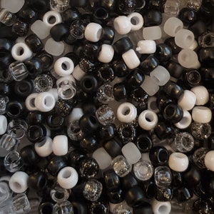 Puede incluir: Una colección de cuentas variadas en negro, blanco y transparente. Las cuentas son de diferentes formas y tamaños, incluyendo redondas, cilíndricas y facetadas. La imagen muestra una vista de cerca de las cuentas, con una mezcla de materiales opacos y translúcidos.