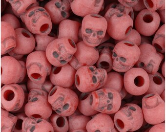 May include: A close-up shot of numerous pink skull-shaped beads. Each bead has a detailed skull design with dark gray accents and a hole running through the center. The beads are scattered, creating a textured, spooky aesthetic.