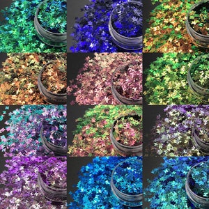 Puede incluir: Una colección de purpurina en forma de mariposa en varios colores, incluyendo verde, azul, dorado y morado. La purpurina está contenida en pequeños botes de plástico transparente, dispuestos en un patrón de cuadrícula. Para manualidades o nail art.