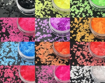 May include: A collection of small, butterfly-shaped craft supplies in various colors, including black, yellow, green, pink, purple, orange, blue, and white. Each color is contained in a clear, round container, arranged in a grid pattern against a black background.