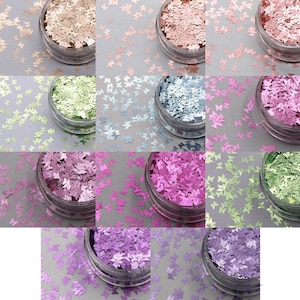 Puede incluir: Una colección de purpurina en forma de mariposa en varios colores, incluyendo dorado, rosa, verde y morado, presentada en botes de plástico transparente. La purpurina está esparcida alrededor de los botes sobre una superficie gris, creando un efecto brillante.