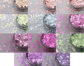 May include: A collection of small, butterfly-shaped glitter in various colors, including gold, pink, green, and purple, displayed in clear plastic jars. The glitter is scattered around the jars on a gray surface, creating a shimmering effect. Ideal for crafts and decorations.