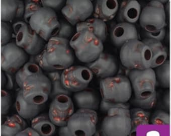 May include: A collection of matte black skull-shaped beads featuring subtle red accents within the recessed details of the eye sockets and nasal cavities. Each bead has a central hole for stringing, creating a uniform pile of dark skull beads.
