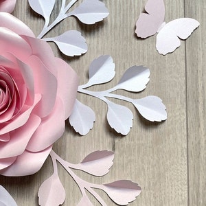 Könnte beinhalten: Rosa und weiße Papierblumen und Blätter auf einem Holzgrund. Die Blumen sind geschichtet und haben einen 3D-Effekt. Die Blätter sind zart und haben einen hellrosa Farbton.