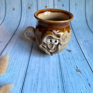 Peut inclure: Une tasse en céramique faite à la main avec un motif de visage souriant. La tasse a une base beige avec un bord et une anse marron. Le visage a des yeux blancs et une bouche souriante. La tasse est sur une surface en bois bleu clair.