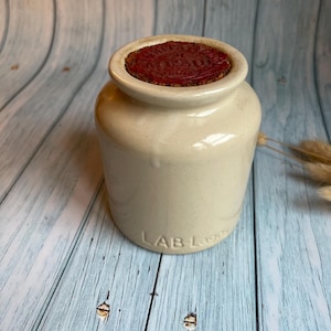 Puede incluir: Un tarro de cerámica blanco con una tapa de corcho roja. El tarro tiene el texto "LAB-L" impreso en la parte inferior.