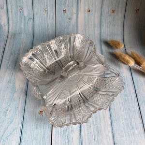 May include: Clear, vintage glass serving dish with a decorative, scalloped edge. The dish has a central handle and is divided into three sections. The glass has a textured, patterned design. The dish is on a light blue wooden surface.