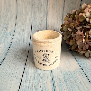 Może przedstawiać: Kremowy ceramiczny garnek z napisem "J. Sainsbury's Freshly Made Bloater Paste" w czarnych literach. Garnek ma cylindryczny kształt i lekko postarzany wygląd. Tło to jasnoniebieska drewniana powierzchnia.