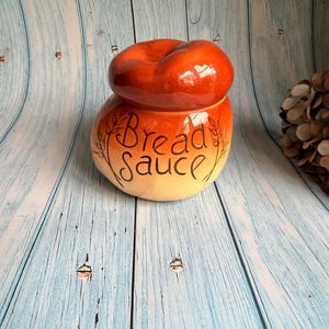 May include: A ceramic bread sauce jar with a lid. The jar is a gradient of orange and cream with the words "Bread Sauce" in black script. The lid is a darker orange, and the jar has a decorative leaf design.