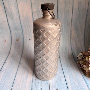 May include: A silver metal canteen with a black cap and a diamond pattern on the body. The canteen has a small metal loop attached to the cap. The background is a light blue wooden surface.