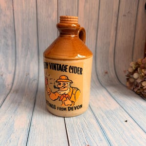 Könnte beinhalten: Vintage Apfelwein-Krug mit braunem Deckel und Griff sowie einem beigefarbenen Körper. Der Krug zeigt eine Cartoon-Illustration eines Mannes mit Pfeife, mit dem Text "Vintage Cider" und "Greetings from Devon".