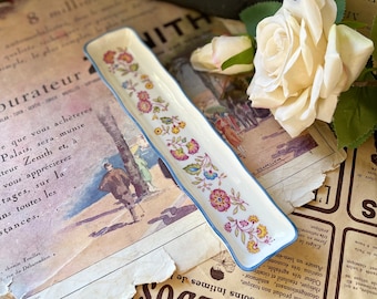 May include: A white rectangular dish with a blue rim and a floral pattern in shades of pink, yellow, and green. The dish is on a surface with printed text.