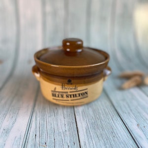 以下が含まれることがあります： 白い文字で「Harrods Blue Stilton」と書かれた蓋付きの茶色の陶器の容器。容器は茶色の釉薬とクリーム色の底面をしています。