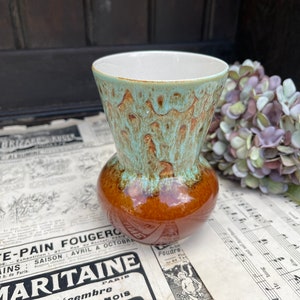 May include: A small, brown and turquoise glazed ceramic vase with a rounded body and a flared opening. The vase has a mottled glaze with a light turquoise color on the top and a brown color on the bottom.