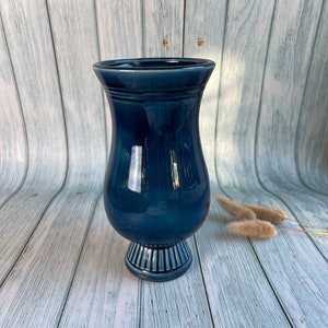 May include: A dark blue ceramic vase with a flared top and a pedestal base. The vase is sitting on a light wood surface.