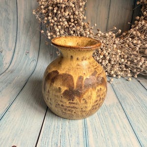 May include: A handcrafted ceramic vase with a rounded body and a flared rim. The vase features a warm, earthy color palette with shades of yellow and brown. The design includes a textured, mottled pattern, adding visual interest to the decorative piece.