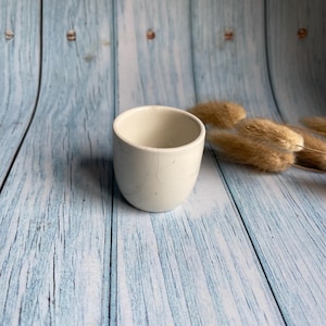 Könnte beinhalten: Eine kleine, weiße Keramiktasse mit leicht abgerundeter Form. Die Tasse steht auf einer hellblauen Holzoberfläche.