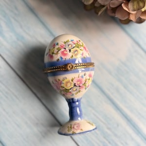May include: An egg-shaped trinket box with a floral design in pink, yellow, and blue. The box is supported by a blue stem and base, and has a gold-colored band around the middle. The egg-shaped box is a decorative item.