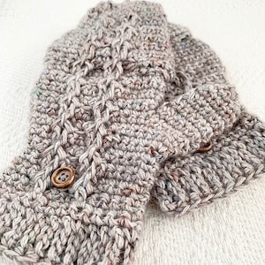 Könnte beinhalten: Ein Paar gestrickte Fingerlose Handschuhe mit einem Zopfmuster und braunen Knöpfen. Die Handschuhe sind aus einem melierten Garn in Grautönen und Braun gefertigt.