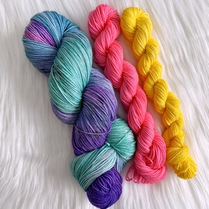 May include: Three skeins of yarn in different colours: a teal, purple, and pink ombre yarn, a bright pink yarn, and a bright yellow yarn.