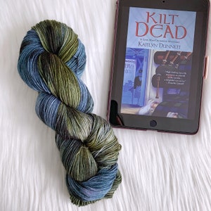 May include: A skein of yarn in shades of green, blue, and black. The yarn is arranged in a loop and is sitting on a white surface. A book titled "Kilt Dead" is visible in the background.