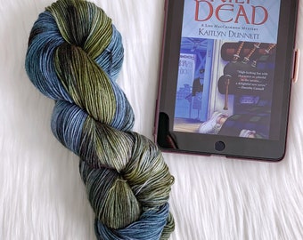 May include: A skein of yarn in shades of green, blue, and black. The yarn is arranged in a loop and is sitting on a white surface. A book titled "Kilt Dead" is visible in the background.