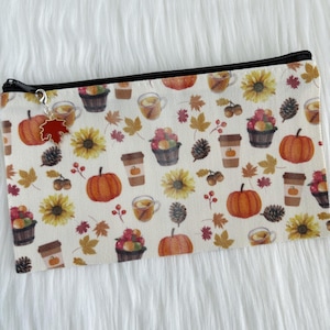 Op de afbeelding: Een rechthoekige etui met een zwarte ritssluiting en een bedel. De etui heeft een herhalend patroon van pompoenen, zonnebloemen, koffiekopjes en herfstbladeren op een crèmekleurige achtergrond. De bedel is een rood blad.