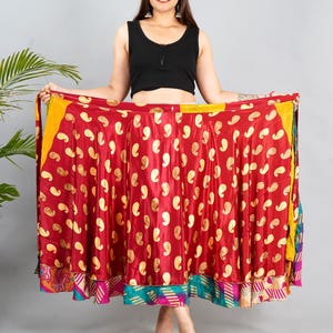 May include: A flowing, red skirt with a gold paisley pattern. The skirt has a layered design with colorful patterned trim. The model is wearing a black top. The skirt is a vibrant piece of clothing.